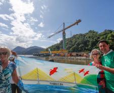 Governador Ratinho Júnior e duas mulheres segurando uma bandeira com a ponte desenhada